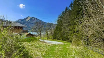 Panoramablick auf ein Haus am Seeufer mit Bergen und Wald im Hintergrund unter blauem Himmel.