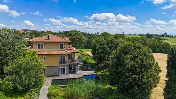 Haus mit Teich und viel Grün in ländlicher Umgebung unter blauem Himmel.