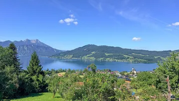 Panoramablick auf den See und die umliegenden Berge mit grüner Landschaft unter blauem Himmel.