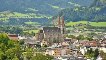 Panoramablick auf eine malerische Stadt mit einer imposanten Kirche, umgeben von grünen Hügeln und Bergen.
