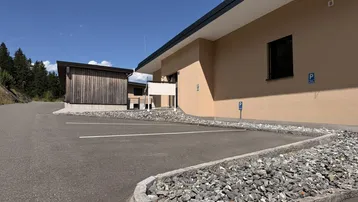 Außenansicht eines Gebäudes mit beiger Fassade und angrenzendem Parkplatzbereich unter blauem Himmel.