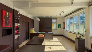 Geräumiges Büro mit großen Fenstern, Holzboden und modernen Möbeln in dunklen Farbtönen, gut beleuchtet.