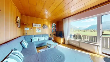 Helles Wohnzimmer mit Holzvertäfelung, großem Sofa und weitem Blick auf die Berge und grüne Landschaft.