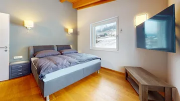 Schlafzimmer mit grauem Bett, Holzboden und Fensterblick auf die verschneite Berglandschaft.