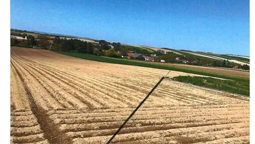 Gepflügtes Feld mit sichtbaren Furchen und einem blauen Himmel, im Hintergrund sind Häuser zu erkennen.