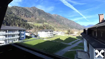 Weitläufiger Blick von einem Balkon auf die umliegende Berglandschaft und Wohngebäude unter blauem Himmel.
