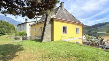 Gelbes Haus mit Satteldach und großem Gartenbereich in bergiger Umgebung.