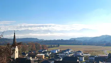 Panoramablick über ein Dorf mit Feldern und einer beeindruckenden Bergkette im Hintergrund unter blauem Himmel.