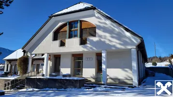 Weißes Gebäude mit Giebeldach und großen Fenstern, umgeben von Schnee an einem sonnigen Wintertag mit Bergblick.