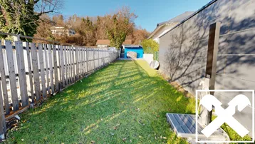 Langer, gepflegter Gartenbereich mit Rasenfläche und Holzzaun, umgeben von Bäumen und einem Geräteschuppen.