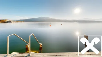 Weitläufiger Seeblick von einem Steg aus, mit klarem Wasser und sonnigem Himmel, umgeben von Bergen.