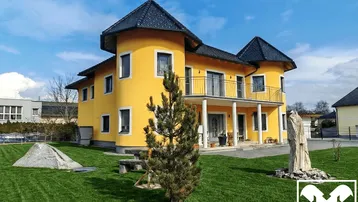 Großzügiges Einfamilienhaus mit gelber Fassade, Türmchen und gepflegtem Vorgarten unter blauem Himmel.
