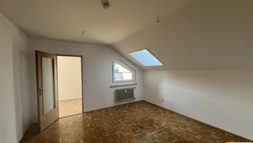 Helles Zimmer im Dachgeschoss mit Parkettboden, Dachfenster und schrägen Wänden.