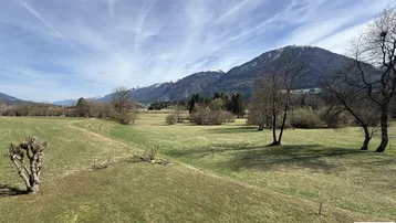 Weitläufige grüne Landschaft mit Bergen im Hintergrund unter einem blauen Himmel und wenigen Wolken.