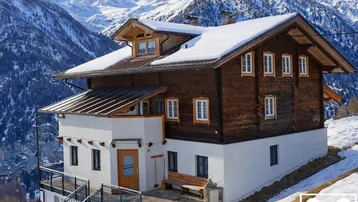 Traditionelles Holzhaus mit weißem Sockel und schneebedecktem Dach in alpiner Berglandschaft.