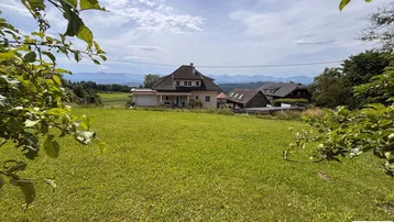 Weitläufige Grünfläche mit Einfamilienhaus im Hintergrund und Bergblick unter bewölktem Himmel.