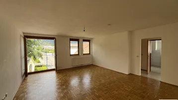 Helles, leeres Wohnzimmer mit Parkettboden und Zugang zu einem kleinen Balkon mit Blick ins Grüne.