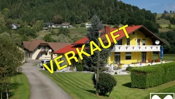 Gelbes Haus mit rotem Satteldach, umgeben von gepflegtem Garten und grüner Hügellandschaft.
