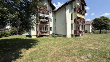 Mehrfamilienhaus mit mehreren Balkonen und großem Rasenbereich vor einer Bergkulisse.
