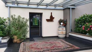 Eingangsbereich eines Gebäudes mit Glasüberdachung, dunklen Fliesen und einem dekorativen Teppich vor der Tür.