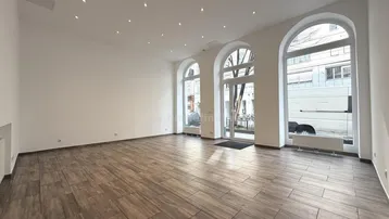 Weitläufiger Raum mit elegantem Holzfliesenboden, weißen Wänden und hohen Rundbogenfenstern zur Straße.