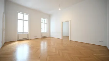 Helles, leeres Zimmer mit Fischgrätparkett und hohen Fenstern, ideal als Wohnbereich nutzbar.