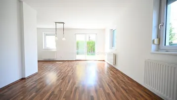 Helles, leeres Wohnzimmer mit Laminatboden und Zugang zum Garten durch eine Schiebetür.