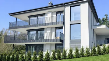Modernes Mehrfamilienhaus mit weißer Fassade, großen Fenstern und Balkonen in grüner Umgebung.