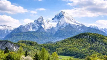 Malerische Berglandschaft mit schneebedeckten Gipfeln und grünen Wäldern unter blauem Himmel.