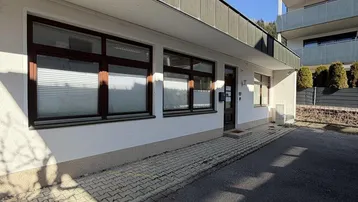 Weißes Geschäftsgebäude mit braunen Fensterrahmen und gepflastertem Eingangsbereich unter blauem Himmel.