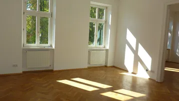 Helles Zimmer mit Fischgrätparkett und großen Fenstern, die viel Tageslicht hereinlassen.