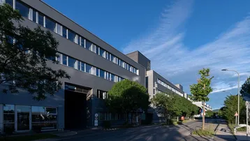 Modernes Bürogebäude mit dunkler Fassade und großen Fenstern entlang einer Straße unter blauem Himmel.