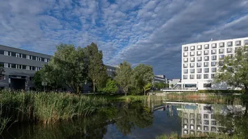 Blick auf moderne Bürogebäude und einen Teich mit Schilf unter einem bewölkten Himmel.