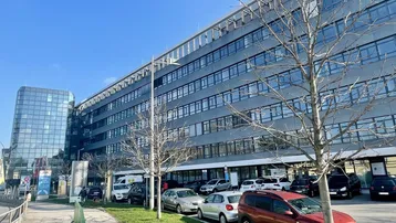 Modernes Bürogebäude mit Glasfassade und Parkplätzen davor unter blauem Himmel.