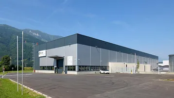 Modernes Industriegebäude mit grauer Fassade und weitem Blick auf die umliegende Berglandschaft unter blauem Himmel.