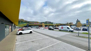 Blick auf den Parkplatz eines Einkaufszentrums mit verschiedenen Geschäften und geparkten Autos unter bewölktem Himmel.