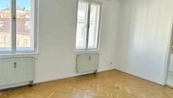 Helles Zimmer mit Fischgrätparkett, zwei großen Fenstern und Blick auf die Umgebung.