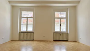 Heller, leerer Raum mit Fischgrätparkett und zwei hohen Fenstern, die viel Tageslicht hereinlassen.