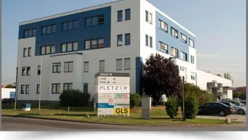 Modernes Bürogebäude mit blau-weißer Fassade und großen Fenstern, umgeben von Grünflächen und einem Firmenschild.