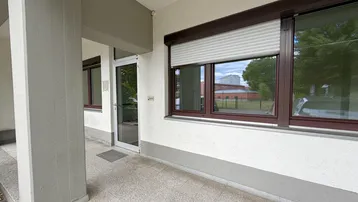 Eingangsbereich eines Gebäudes mit großen Fenstern, Rollläden und einer gepflasterten Fläche.