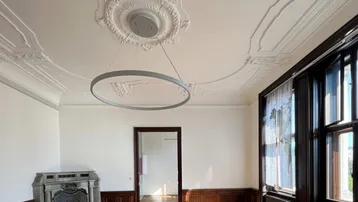 Geräumiges Wohnzimmer mit Fischgrätparkett, hohen Decken, Stuckverzierungen und einem historischen Kamin.