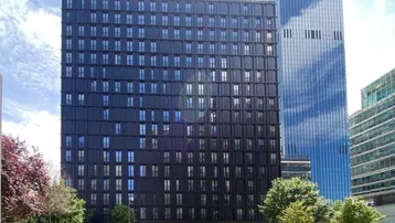 Modernes, dunkles Hochhaus mit vielen Fenstern und einer gepflegten Grünanlage davor, unter blauem Himmel.