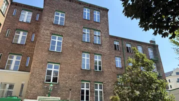 Historisches Backsteingebäude mit vielen weißen Fenstern und grünen Akzenten unter blauem Himmel.