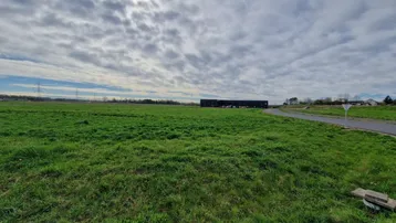 Weitläufiges grünes Feld unter bewölktem Himmel mit Industriegebäuden in der Ferne.