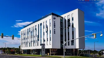 Modernes Bürogebäude mit weißer Fassade und großen Fenstern an einer belebten Kreuzung unter blauem Himmel.