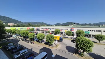 Luftaufnahme eines modernen Gewerbegebiets mit großem Parkplatz und Bergen im Hintergrund unter blauem Himmel.