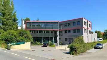 Modernes Bürogebäude mit großen Fenstern und Parkplätzen im Freien, umgeben von Bäumen und Grünflächen.