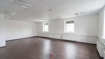 Geräumiges, leeres Büro mit dunklem Laminatboden, weißen Wänden und zwei Fenstern mit Jalousien.