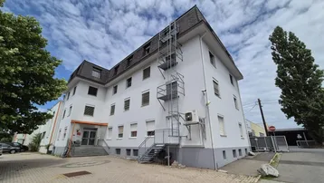 Mehrstöckiges Gebäude mit weißer Fassade, dunklem Dach und externer Metalltreppe unter bewölktem Himmel.