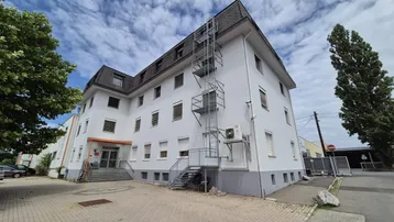Weißes Geschäftsgebäude mit Mansarddach, externer Fluchttreppe und Klimaanlage an der Fassade.